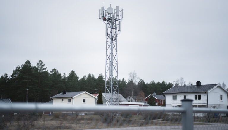 Korkea matkapuhelinmasto seisoo omakotitalojen ja mäntyjen takana pilvisenä päivänä aidan takaa kuvattuna.